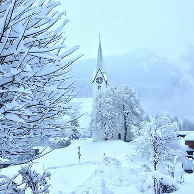 Eine verschneite Landschaft mit einer Kirche im Hintergrund. Bäume und Sträucher sind vollständig mit Schnee bedeckt.