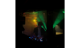 Ein DJ steht hinter einem Mischpult in einem dunklen Raum. Bunte Lichtstrahlen schaffen eine lebendige Atmosphäre.
