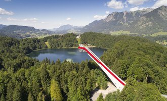 Eine beeindruckende Aussichtsbrücke über einen klaren See, umgeben von grünen Wäldern und Bergen. Der Himmel ist blau mit einigen Wolken.