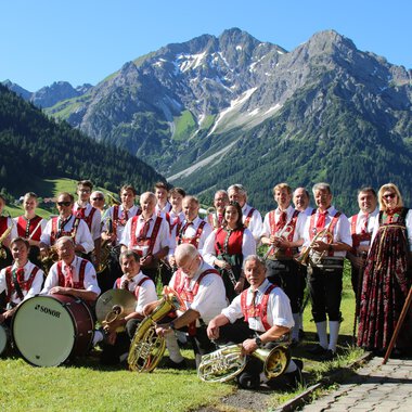 A group of musicians in traditional attire stands in front of a mountainous landscape. They are holding musical instruments and a flag in their hands. | © Musikverein d´Hirschegger | Jockl Metzger