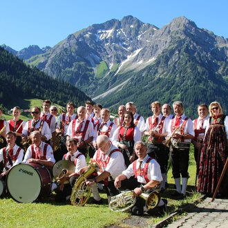 Eine Gruppe von Musikern in traditioneller Tracht steht vor einer bergigen Landschaft. Sie halten Musikinstrumente und eine Fahne in der Hand. | © Musikverein d´Hirschegger | Jockl Metzger