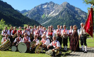 Eine Gruppe von Musikern in traditioneller Tracht steht vor einer bergigen Landschaft. Sie halten Musikinstrumente und eine Fahne in der Hand. | © Musikverein d´Hirschegger | Jockl Metzger