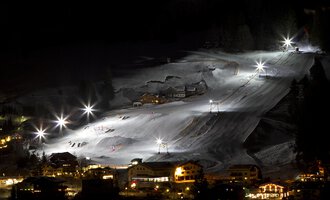 A beautifully lit ski resort at night with white snow-covered mountains. The lifts and slopes are clearly visible, while the surrounding buildings are gently illuminated.