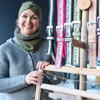 A smiling woman stands in front of a stand with colorful ribbons. She is wearing a gray sweater and a scarf.