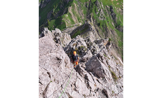 An impressive mountain landscape with steep cliffs and green meadows. A climber is on a rock face. | © stoamandl.at | Olli Sinz