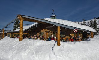 Eine gemütliche Berghütte im Schnee mit Gästen auf der Terrasse. Der Himmel ist klar und blau, und die Umgebung ist winterlich. | © Bergstüble | Michaela Beck Dornach