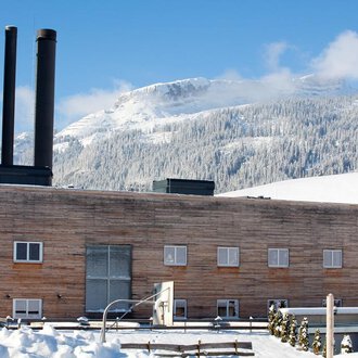 Ein modernes Gebäude mit Holzfassade und drei Schornsteinen in einer schneebedeckten Landschaft. Im Hintergrund sind Berge und ein klarer blauer Himmel zu sehen.