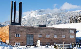 Ein modernes Gebäude mit Holzfassade und drei Schornsteinen in einer schneebedeckten Landschaft. Im Hintergrund sind Berge und ein klarer blauer Himmel zu sehen.