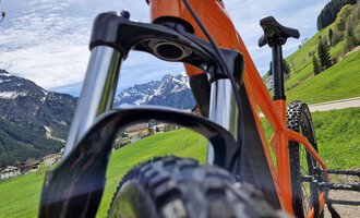 Ein orangefarbenes Mountainbike in einer grünen Landschaft mit Bergen im Hintergrund. Der Himmel ist leicht bewölkt und es ist eine idyllische Umgebung. | © Austrian Ski- & Service Ranch | Trixi Leitner