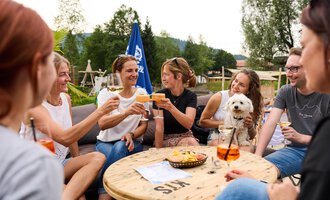 A group of women and men are sitting together at a table and raising glasses for a toast. A small dog is sitting on the table. | © Kleinwalsertal Tourismus | Oliver Farys