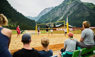 A beach volleyball game in the mountains with a group of spectators. In the background, green forests and majestic mountains are visible. | © Kleinwalsertal Tourismus | Oliver Farys
