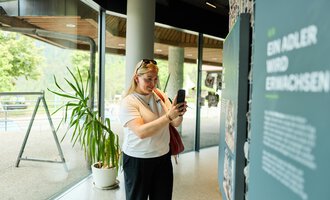A visitor is photographing an exhibition panel in a modern room. Behind her is a large plant, and there are windows with a view of nature. | © Kleinwalsertal Tourismus | Oliver Farys