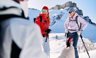 Two women are standing in the snow in the mountains and laughing. They are wearing ski clothing and holding hiking sticks. | © Kleinwalsertal Tourismus | Oliver Farys