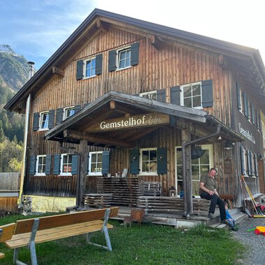 Ein traditionelles Holzhaus mit dem Namen "Gemstelhof" steht in einer ländlichen Umgebung. Vor dem Gebäude sitzen Bänke und ein Mensch ist sichtbar, während die Berge im Hintergrund zu sehen sind. | © Bernhards Gemstel Hofladen | Bernhard Heim