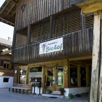 A wooden building with a sign that says "BioHof." Next to it are tables and chairs in the outdoor area, surrounded by other traditional buildings. | © Kleinwalsertal Tourismus