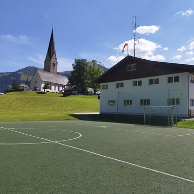 A football pitch in front of a modern building with a church in the background. The surroundings are green with hills and a clear blue sky. | © Kleinwalsertal Tourismus | N. Lughammer