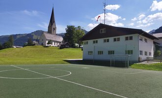 Ein Fußballplatz vor einem modernen Gebäude mit einer Kirche im Hintergrund. Die Umgebung ist grün mit Hügeln und einem klaren blauen Himmel. | © Kleinwalsertal Tourismus | N. Lughammer