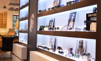 A stylish room with a presentation of sunglasses on illuminated shelves. In the background, there are images of models wearing glasses. | © Bräuer Optik & Akustik | AlpenMedia-Design