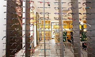 A modern eyewear store with a large selection of eyeglass frames. The wall is adorned with various glasses on display, while a promotional poster can be seen in the background. | © Bräuer Optik & Akustik | AlpenMedia-Design