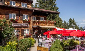 Ein charmantes Gasthaus mit Holzfassade und blühenden Blumen. Im Freien sind Tische mit roten Sonnenschirmen aufgestellt. | © Bühlalpe | Frank Drechsel