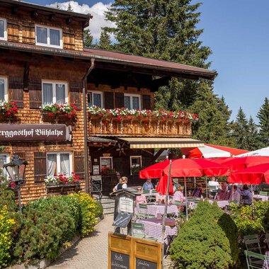 Ein charmantes Gasthaus mit Holzfassade und blühenden Blumen. Im Freien sind Tische mit roten Sonnenschirmen aufgestellt. | © Bühlalpe | Frank Drechsel