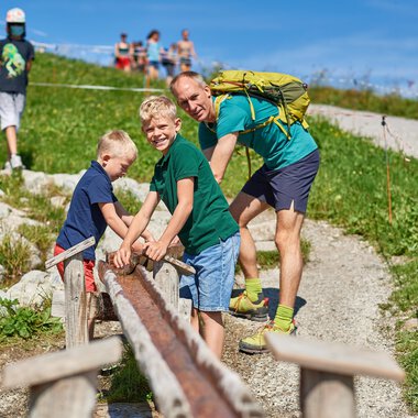Vater und seine zwei Söhne spielen auf einem Weg im Freien. Im Hintergrund sind weitere Personen zu sehen, und die Umgebung ist grün und sonnig. | © OBERSTDORF · KLEINWALSERTAL BERGBAHNEN