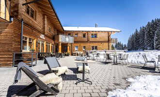 A cozy wooden building in winter with a terrace.  
Chairs and tables are on the snow-covered ground, surrounded by snow-covered trees.