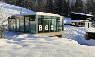 Ein modernes, gläsernes Gebäude mit der Aufschrift "BOX" steht inmitten einer schneebedeckten Landschaft. Im Hintergrund sind schneebedeckte Bäume und weitere Gebäude sichtbar. | © Sport Kessler GmbH