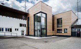 A modern building with a wooden façade and large glass windows. It is located in a rural area with other traditional buildings. | © Feuerwehr Riezlern | Gemeinde Mittelberg