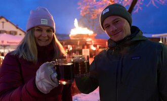 Two people are clinking glasses and smiling. In the background, a festive atmosphere with a fire pit and seasonal lights can be seen.