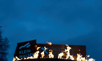 A sign with the inscription "Klein Walsertal" is burning in front of a dark sky. The flames glow brightly and create a dramatic atmosphere.
