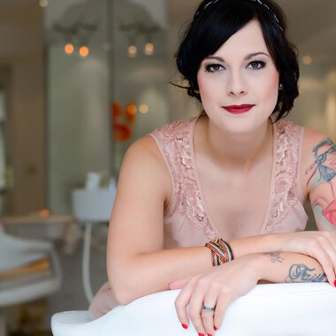 Eine Frau mit Tattoos sitzt lächelnd in einem modernen Raum. Sie trägt ein elegantes, rosafarbenes Kleid und hat einen schicken Look. | © Fräulein Sugar Coiffeur | Sandra Bromm