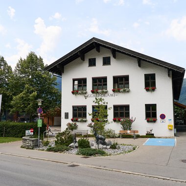 A traditional two-story house with decorative windows and a clear sky. Surrounded by trees and a quiet street. | © Kleinwalsertal Tourismus | Oliver Farys
