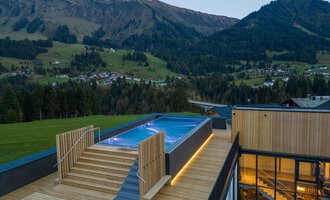 Ein modernes Gebäude mit einem Holzdeck und einem Pool, der in die Landschaft eingebettet ist. Im Hintergrund sind die Berge und die umliegende Natur sichtbar.