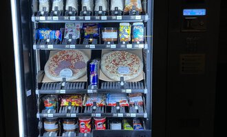 Ein Snackautomat mit verschiedenen Getränken und Lebensmitteln. Darunter befinden sich Pizzen, Süßigkeiten und Energydrinks.