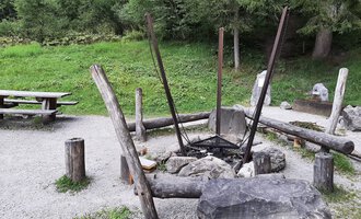 A cozy outdoor grill area with a large fire circle made of stones and wood. In the background, there is a picnic bench on a green meadow. | © Kleinwalsertal Tourismus | Veronika Senn