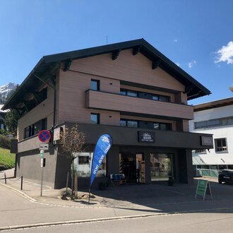 Ein modernes Gebäude mit mehreren Etagen liegt an einer ruhigen Straße. Im Hintergrund sind Berge und weitere Gebäude zu sehen. | © Sport Hilbrand GmbH