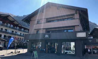 Ein modernes Gebäude mit einem Geschäft im Erdgeschoss. Die Sonne scheint auf die Fassade, und im Hintergrund sind weitere Häuser und Berge zu sehen. | © Sport Hilbrand GmbH