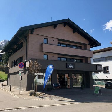 A modern building with several floors is located on a quiet street. In the background, mountains and additional buildings can be seen. | © Sport Hilbrand GmbH