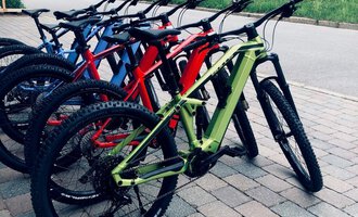 Eine Reihe von bunten Mountainbikes steht auf einem gepflasterten Weg. Im Hintergrund ist ein Holzhaus und eine grüne Wiese zu sehen. | © Sport Hilbrand GmbH