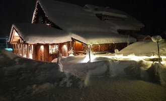 Ein gemütliches Haus im Schnee bei Nacht, beleuchtet von warmem Licht. Der Schnee bedeckt die Umgebung und schafft eine winterliche Atmosphäre.