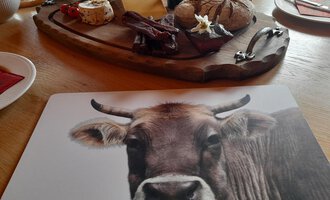 Ein rustikales Holzbrett mit verschiedenen Käsesorten, Wurst und frischem Brot. Der Tisch ist gemütlich gedeckt und im Vordergrund ist ein Platzset mit dem Bild einer Kuh zu sehen.