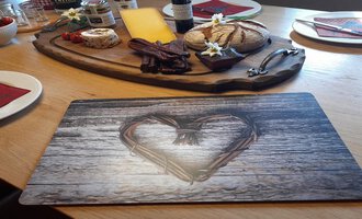 Ein gedeckter Tisch mit einer Holzplatte voller Käse, Wurst und Brot. Im Vordergrund liegt eine Platzmatte mit einem Herz-Muster.