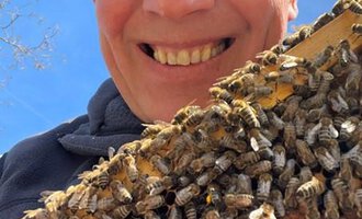Ein Mann lächelt vor einer Bienenwabe, die mit vielen Bienen bedeckt ist. Der Himmel ist blau und sonnig. | © Imkerei Jürgen Scharnagl