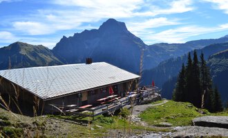 Eine alpine Hütte umgeben von Bergen und Wäldern. Der Himmel ist klar mit ein paar Wolken. | © Kleinwalsertal Tourismus