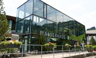Ein modernes Gebäude namens Walserhaus in einer bergigen Umgebung. Umgeben von Bäumen und unter einem klaren Himmel. | © Kleinwalsertal Tourismus | Oliver Farys
