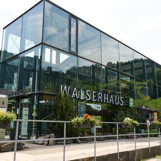 A modern building called Walserhaus in a mountainous environment. Surrounded by trees and under a clear sky. | © Kleinwalsertal Tourismus | Oliver Farys