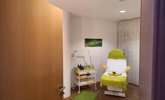 A bright, inviting room with a treatment chair and a small table for cosmetic products. The floor is made of wood and a green carpet creates a relaxed atmosphere. | © Schönzeit | Sebastian Riezler