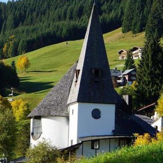 Ein charmantes, modernes Gebäude mit einem hohen spitzen Dach steht inmitten einer grünen Landschaft. Umgeben von Bäumen und sanften Hügeln strahlt die Szene Ruhe und Schönheit aus. | © Ev. Kirche Kleinwalsertal