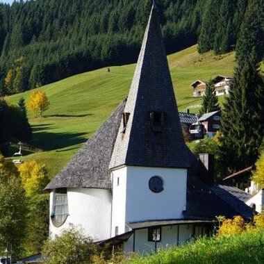 A charming, modern building with a tall pointed roof stands amidst a green landscape. Surrounded by trees and gentle hills, the scene radiates tranquility and beauty. | © Ev. Kirche Kleinwalsertal